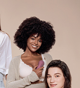 Two women wearing baby carriers with infants. One carrier is light gray, the other is not fully visible. Both women are smiling.