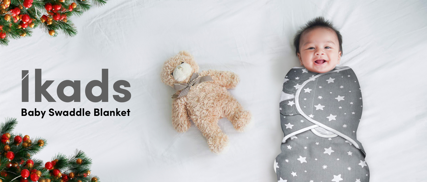 Ikads baby swaddle blanket with star pattern. Gray blanket wrapping a smiling baby, alongside a plush teddy bear. Christmas decorations visible.