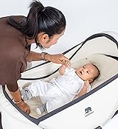 Portable infant bed or travel crib being set up by an adult. Collapsible design with mesh sides visible in a home setting.
