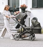 Double stroller with two seats in use outdoors. A woman is bending over the stroller, which contains two young children.