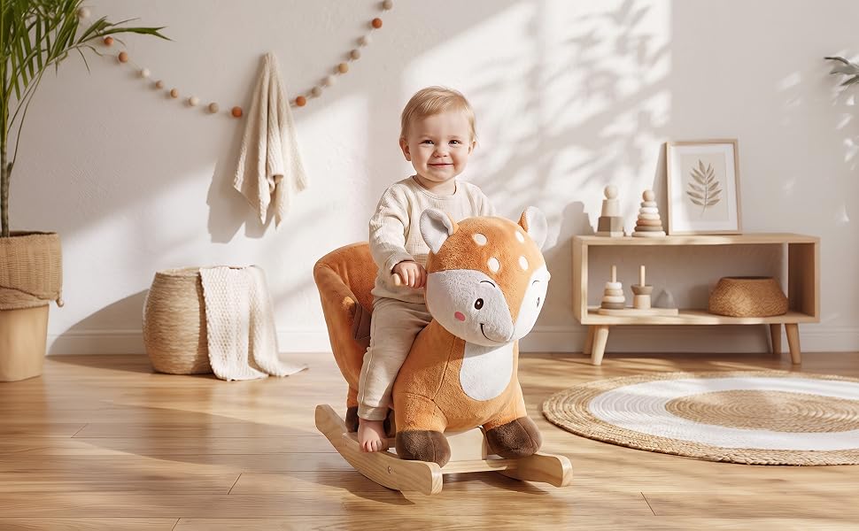 Toddler sitting on wooden floor in bright room with cream-colored plush teddy bear toy. Shelving and woven baskets visible in background.