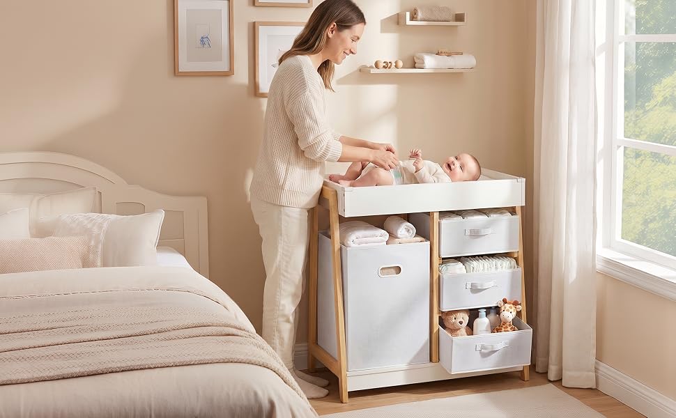 Woman in cream cardigan organizing items in a white bedside storage unit with drawers and fabric compartments next to a bed with white bedding in a bedroom setting.